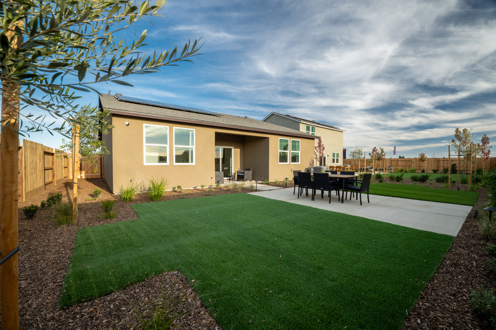 A house with a lawn and a fence and a yard with a table and chairs and a tree.